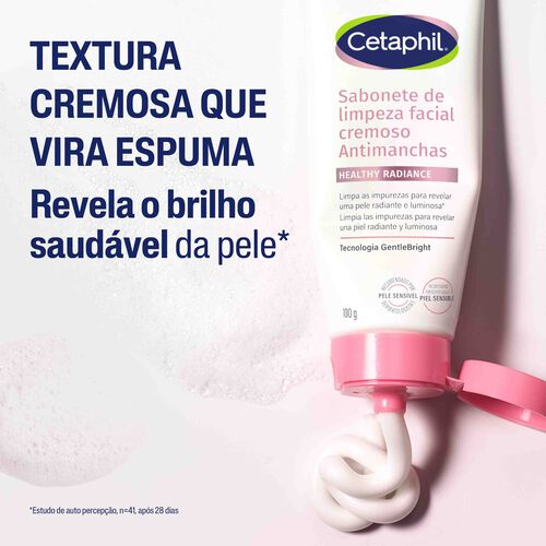 Imagem de um tubo de sabonete de limpeza facial cremoso antimanchas Cetaphil Healthy Radiance. O produto está sendo dispensado, mostrando sua textura cremosa que se transforma em espuma. O texto destaca que o produto revela o brilho saudável da pele.