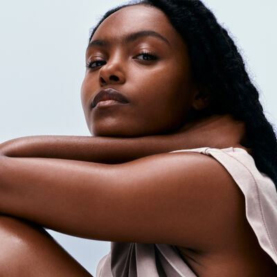 Retrato de uma mulher negra com pele radiante e saudável. Ela tem cabelos longos e trançados e está usando uma blusa de alças clara. Seus braços estão cruzados e ela olha diretamente para a câmera com uma expressão serena.