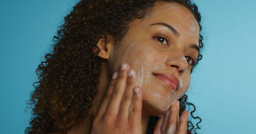 A imagem mostra uma mulher de pele com acne sorrindo enquanto lava o rosto. Ela tem pele morena e cabelos cacheados escuros. Seus dedos estão em contato com a pele do rosto, espalhando um produto de limpeza facial branco e espumoso. O fundo é azul claro, o que cria um contraste com a pele e o cabelo da mulher. A iluminação é suave e uniforme, destacando a textura da pele e a espuma do produto de limpeza.