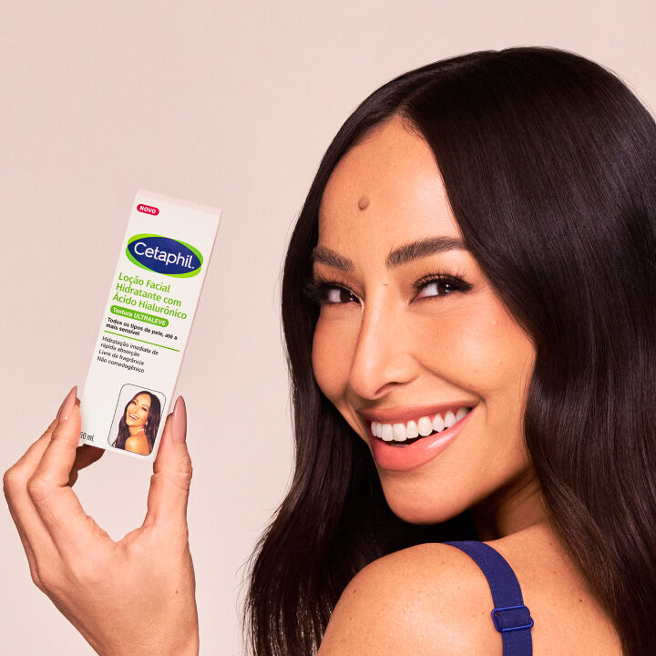 Sabrina Sato sorrindo radiantemente, com seus longos cabelos escuros caindo sobre os ombros, enquanto segura a embalagem da Lo&ccedil;&atilde;o Hidratante Facial com &Aacute;cido Hialur&ocirc;nico Cetaphil. A imagem destaca a import&acirc;ncia da hidrata&ccedil;&atilde;o para uma pele radiante e preparada para a maquiagem.