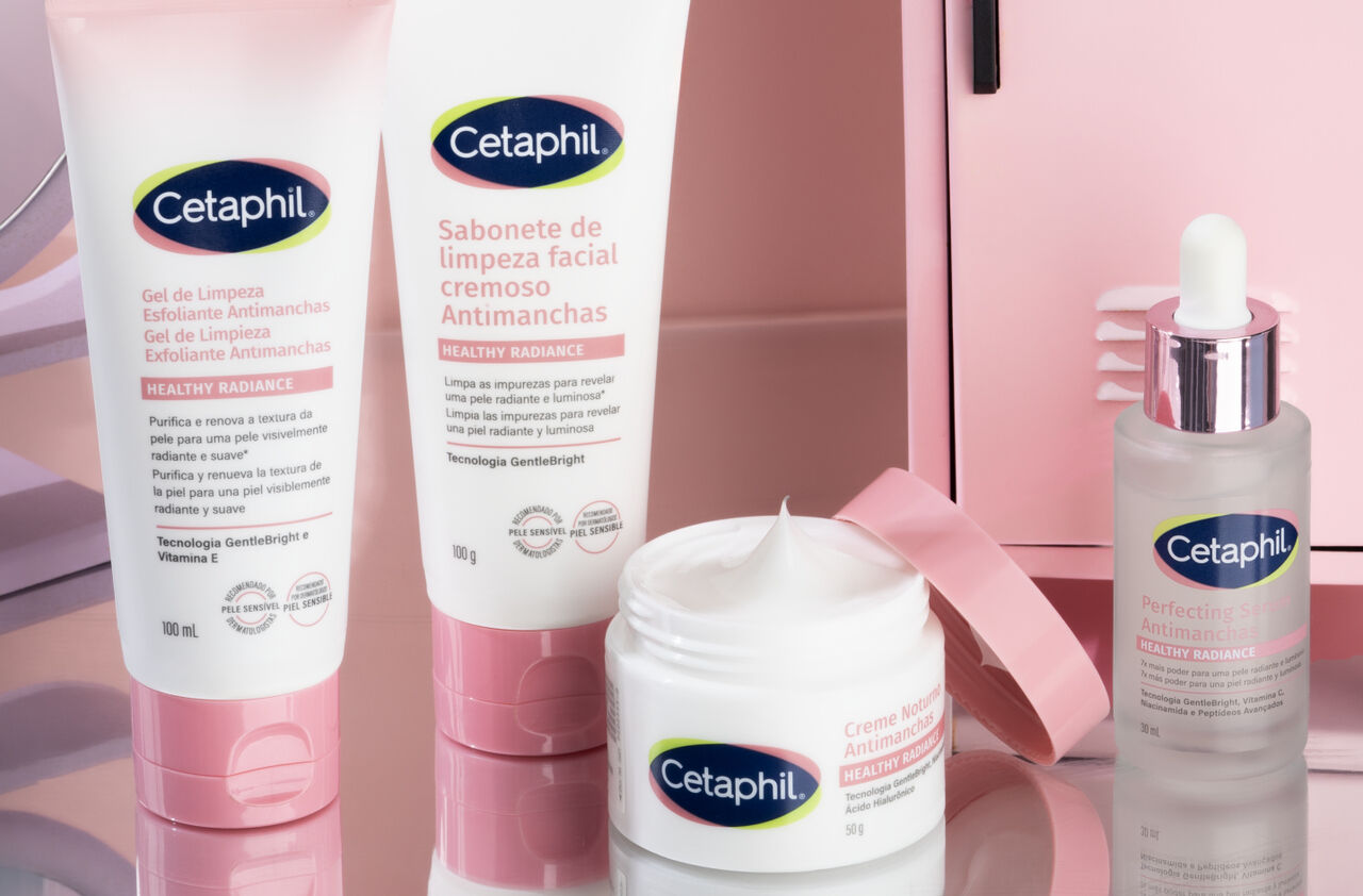 Uma imagem de estúdio exibe quatro produtos da linha Cetaphil "Healthy Radiance" sobre uma superfície rosa-claro reflexiva. Da esquerda para a direita, há dois tubos brancos com detalhes em rosa, um pote branco de creme com uma espátula rosa ao lado, e um frasco transparente com conta-gotas. Todos os produtos têm o logotipo azul e verde da Cetaphil e texto em português, indicando "Gel de Limpeza Esfoliante Antimanchas", "Sabonete de Limpeza Facial Cremoso Antimanchas", "Creme Noturno Antimanchas" e "Sérum Iluminador Antimanchas".