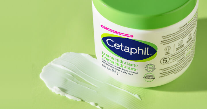 Pote branco Creme Hidratante Cetaphil com tampa verde sobre fundo verde claro, mostrando textura cremosa do produto ao lado. Indicado para pele muito ressecada.