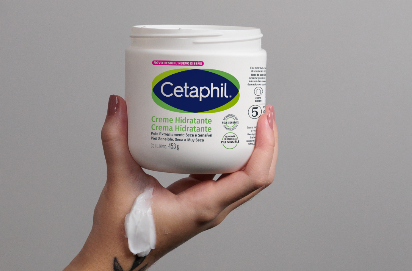 Uma mão com pele escura e unhas pintadas de rosa claro está pegando uma porção de creme branco e espesso do pote de Creme Hidratante Cetaphil. O creme dentro do pote tem uma textura rica e ondulada, indicando sua consistência. O fundo é um tom de cinza-azulado suave e desfocado.