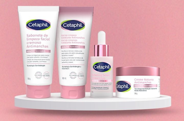 A imagem mostra quatro produtos Cetaphil Healthy Radiance. Da esquerda para a direita, temos o Sabonete de Limpeza Facial, o Gel Esfoliante, o Sérum e o Creme Noturno. Esses produtos estão colocados em cima de uma prateleira circular branca flutuante e a imagem possui um fundo rosa.