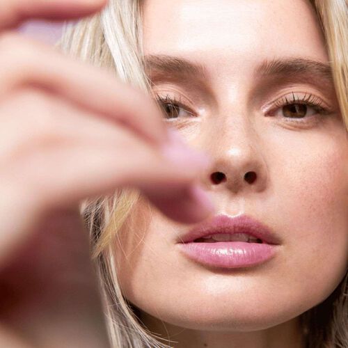 A imagem mostra uma mulher de pele clara e cabelos loiros lisos aplicando um produto branco e iluminado em sua bochecha direita com sua mão direita. Suas unhas estão bem cuidadas, pintadas em tom nude. O fundo em tons suaves de rosa cria uma atmosfera leve, destacando cuidado pessoal e sofisticação.