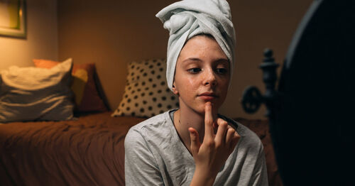 Adolescente branca com toalha em sua cabeça aplicando produto de skincare no rosto e olhando para um espelho. Adolescente branca com toalha em sua cabeça aplicando produto de skincare no rosto e olhando para um espelho.