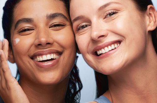 À esquerda, mulher negra sorrindo e usando gel hidratante facial no rosto. À direita, mulher branca sorrindo.