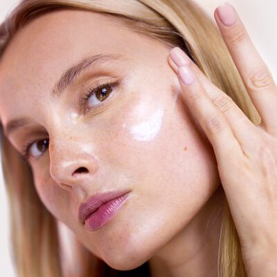 Close-up do rosto de uma mulher branca de olhos castanhos claros com uma pele iluminada e um hidratante na pele. 