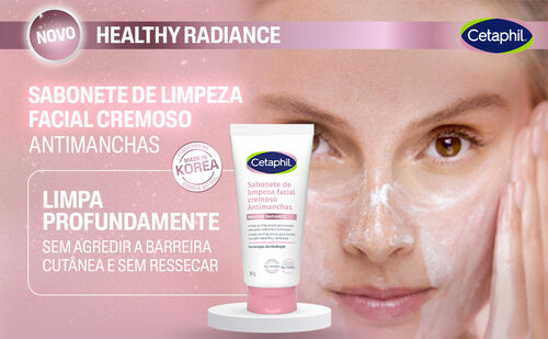 Uma mulher com cabelo escuro e pele clara, com espuma branca de sabonete no rosto, olha para a c&acirc;mera. Sua m&atilde;o direita est&aacute; levantada, com os dedos espalhados. No centro da imagem, um tubo de sabonete de limpeza facial cremoso branco e rosa da Cetaphil est&aacute; em destaque sobre um pedestal branco. O texto "NOVO HEALTHY RADIANCE" e "SABONETE DE LIMPEZA FACIAL CREMOSO ANTIMANCHAS" aparece na parte superior. H&aacute; tamb&eacute;m um selo "MADE IN KOREA" e a frase "LIMPA PROFUNDAMENTE SEM AGREDIR A BARREIRA CUT&Acirc;NEA E SEM RESSECAR".
